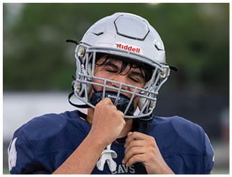A person wearing a white football helmet with a Riddell logo is shown against a blurred outdoor background. Descripci n generada con IA