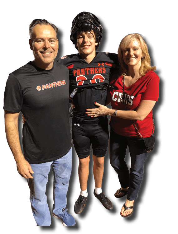 Three people stand together on a sports field at night with one person in a football uniform and helmet, while the others wear casual clothing with matching team logos. Description generated by AI