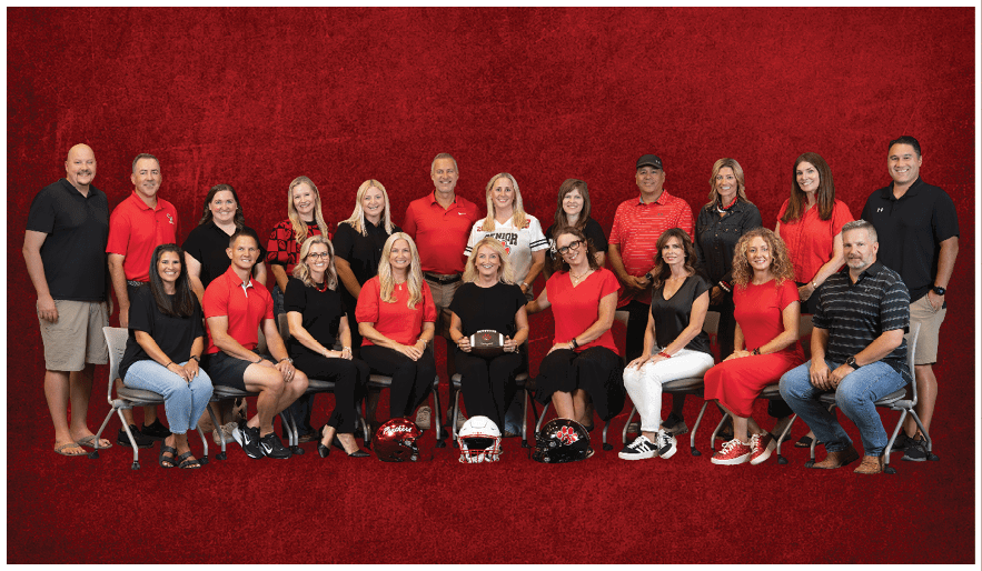 A group of adults dressed mostly in red and black clothing are posed in two rows against a red textured background with three football helmets placed in front of the seated row. Description generated by AI