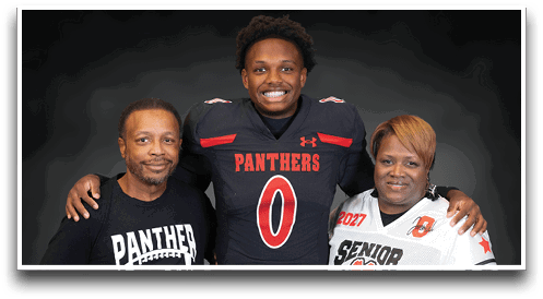 Three people stand together smiling with one wearing a football uniform, another wearing a “Senior Mom" jersey holding a football, and the third in a white button-up shirt. Description generated by AI