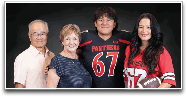 Four adults stand closely together smiling, with two wearing matching football jerseys numbered 67 and holding a football. Description generated by AI