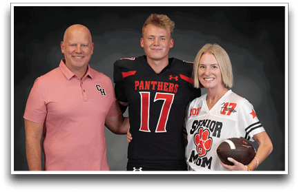 Three adults stand together smiling with one wearing a black sports jersey with the number 17 and the word Panthers, another in a pink polo shirt, and the third in a white sports jersey holding a football with the words Senior Mom. Description generated by AI