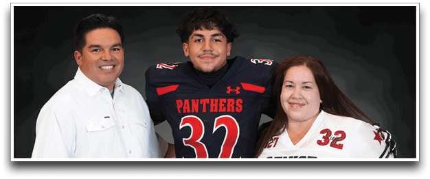 Three people stand closely together smiling, with one person in the center wearing a black football jersey with the number 32 and the word Panthers, while the other two wear casual and sports attire. Description generated by AI