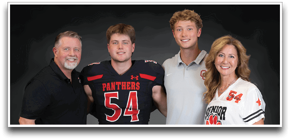 Four people stand closely together smiling against a dark background, with one person wearing a football uniform numbered 54, another holding a football, and the others dressed in casual and sports-themed clothing. Description generated by AI