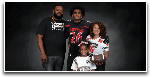 A group of four people, including two wearing football jerseys and one holding a football, pose together against a dark background. Description generated by AI