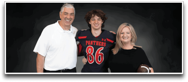 Three people stand together smiling against a dark background with one person in a football uniform and the other two in casual clothing, one holding a football. Description generated by AI