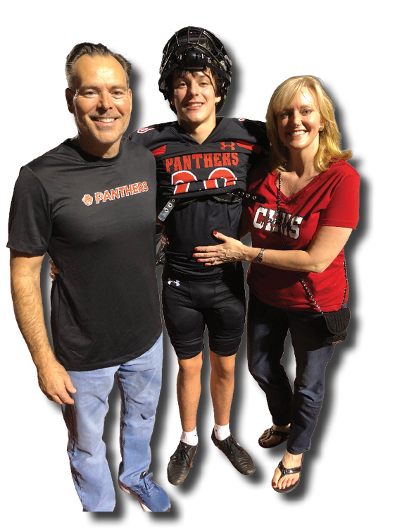 Three people stand together on a sports field at night with one person in a football uniform and helmet, while the others wear casual clothing with matching team logos. Description generated by AI