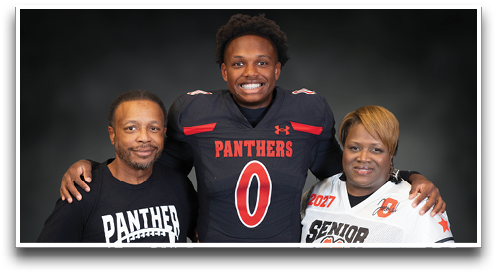 Three people stand together smiling with one wearing a football uniform, another wearing a “Senior Mom" jersey holding a football, and the third in a white button-up shirt. Description generated by AI