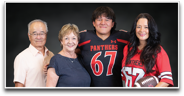 Four adults stand closely together smiling, with two wearing matching football jerseys numbered 67 and holding a football. Description generated by AI