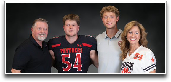 Four people stand closely together smiling against a dark background, with one person wearing a football uniform numbered 54, another holding a football, and the others dressed in casual and sports-themed clothing. Description generated by AI