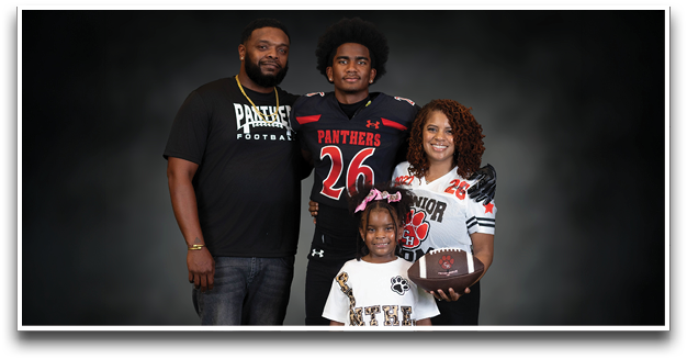 A group of four people, including two wearing football jerseys and one holding a football, pose together against a dark background. Description generated by AI