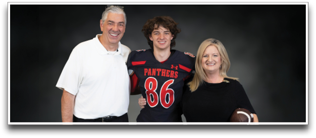 Three people stand together smiling against a dark background with one person in a football uniform and the other two in casual clothing, one holding a football. Description generated by AI