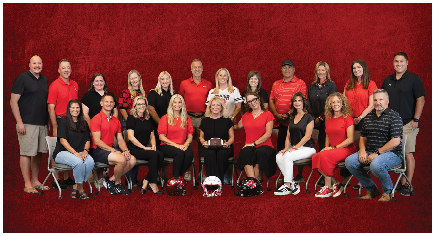 A group of adults dressed mostly in red and black clothing are posed in two rows against a red textured background with three football helmets placed in front of the seated row. Description generated by AI
