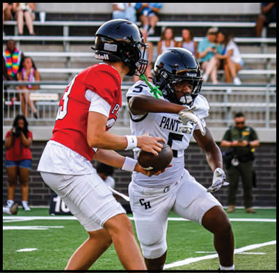 Two people wearing football helmets and uniforms are engaged in a play on a football field with spectators in the background. Description generated by AI