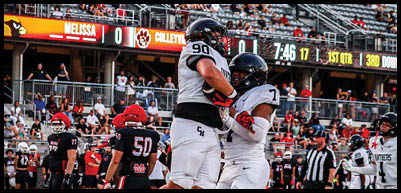 Two athletes in white uniforms celebrate by embracing on a football field while other players and spectators are visible in the background. Description generated by AI