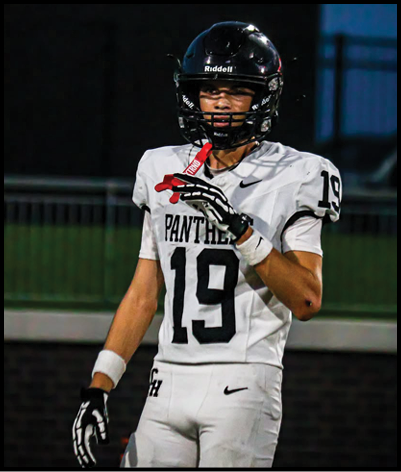 A person wearing a white football jersey with the number 19 and a black helmet is standing on a field at dusk. Description generated by AI