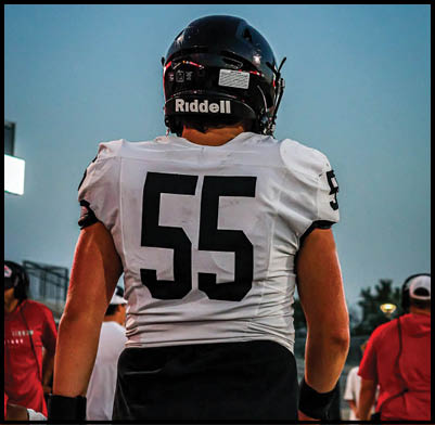 An adult wearing a white football jersey with the number 55 on the back stands on a field with other people in the background. Description generated by AI