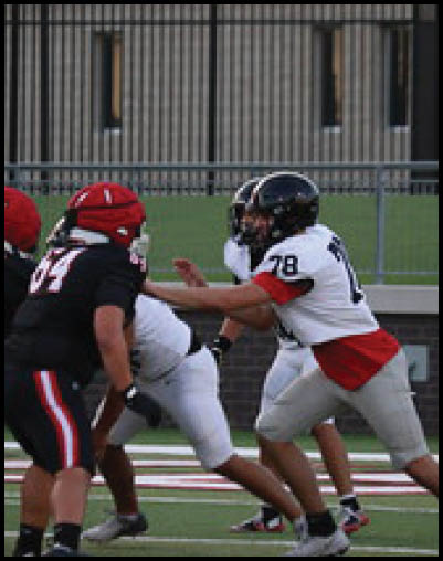 Two people in football uniforms are engaged in a play on a field with a fence and building in the background. Description generated by AI