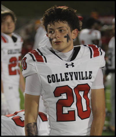 A person wearing a white football uniform with red and black accents and the number 20 stands on a field with other players in the background. Description generated by AI