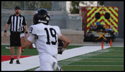 A person wearing a black and white striped referee shirt stands near a person in a football helmet with trees and an emergency vehicle in the background. Description generated by AI
