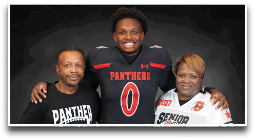 Three people stand together smiling with one wearing a football uniform, another wearing a “Senior Mom" jersey holding a football, and the third in a white button-up shirt. Description generated by AI