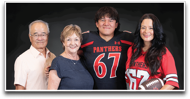 Four adults stand closely together smiling, with two wearing matching football jerseys numbered 67 and holding a football. Description generated by AI