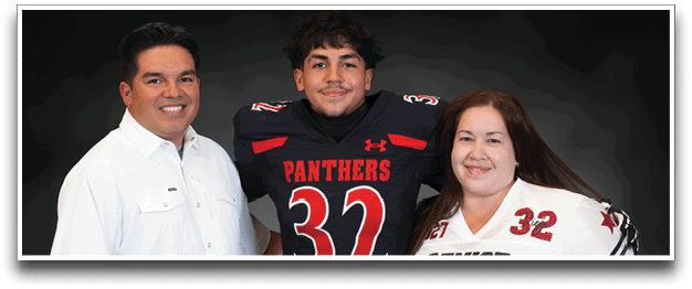 Three people stand closely together smiling, with one person in the center wearing a black football jersey with the number 32 and the word Panthers, while the other two wear casual and sports attire. Description generated by AI