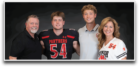 Four people stand closely together smiling against a dark background, with one person wearing a football uniform numbered 54, another holding a football, and the others dressed in casual and sports-themed clothing. Description generated by AI