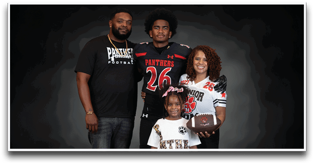 A group of four people, including two wearing football jerseys and one holding a football, pose together against a dark background. Description generated by AI