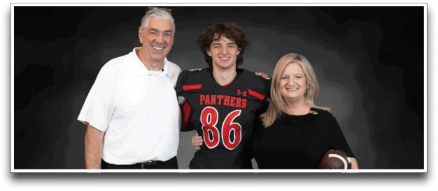 Three people stand together smiling against a dark background with one person in a football uniform and the other two in casual clothing, one holding a football. Description generated by AI
