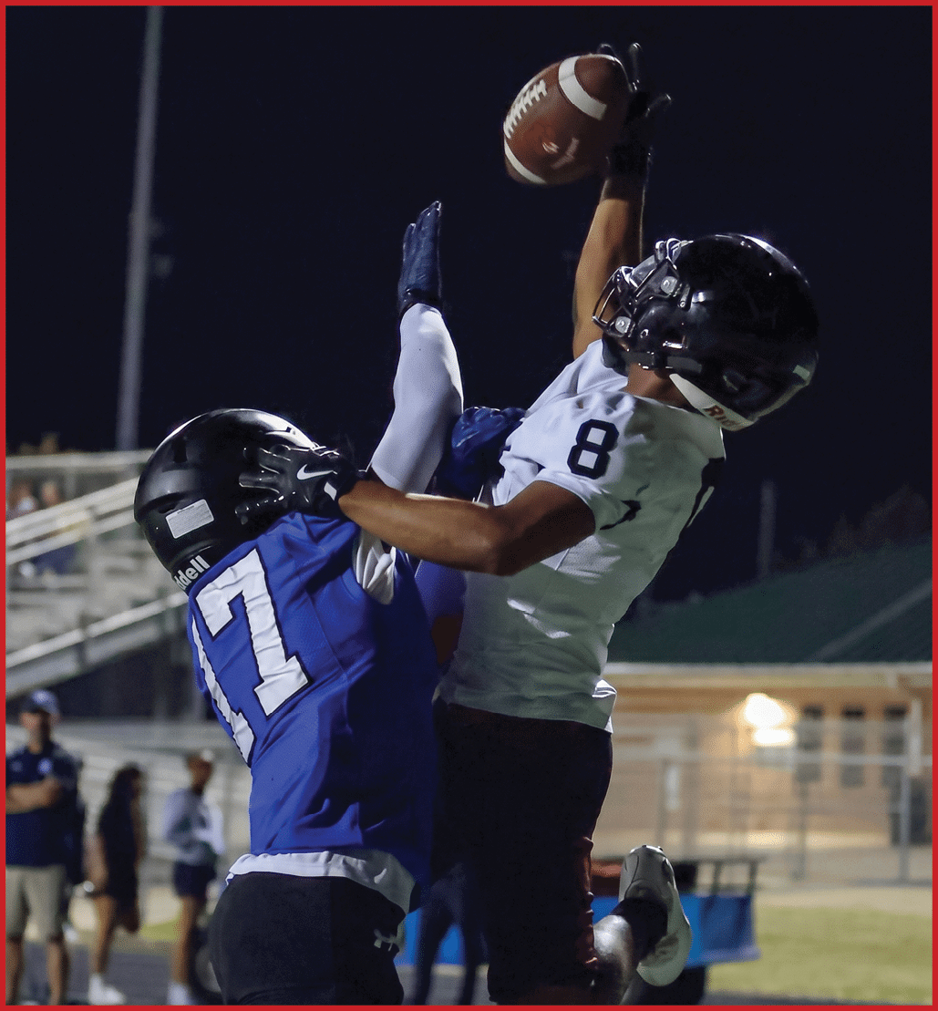 Two people in football gear jump and reach for a football during a nighttime game on a field. Description generated by AI