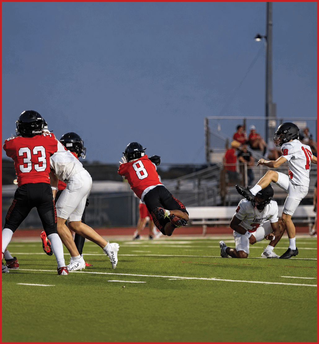 People wearing helmets and sports gear are playing football on a field with a clear sky and spectators sitting on bleachers in the background. Description generated by AI