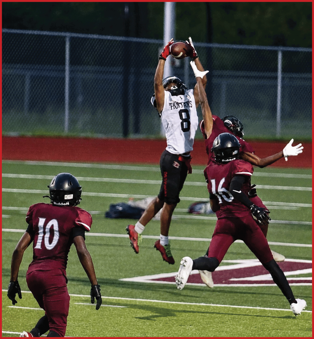 A person wearing a white jersey with the number 8 and the word Trojans is jumping to catch a football while two people in maroon jerseys attempt to block the catch on a football field. Description generated by AI