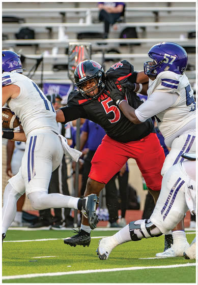 A person in a black and red football uniform is attempting to tackle a person in a white and purple football uniform who is running with the ball while another person in white and purple is blocking. Description generated by AI