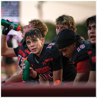 Several people wearing black and red sports uniforms are sitting closely together on a bench, with some drinking from water bottles. Description generated by AI