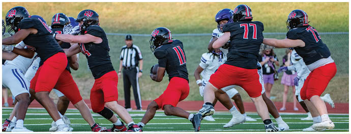 People wearing black helmets, black jerseys, and red pants are playing American football on a field with a referee and spectators in the background. Description generated by AI