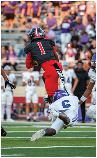 A person in red and black athletic gear is jumping while holding a football as another person in white and purple sports gear attempts to tackle them on a field. Description generated by AI