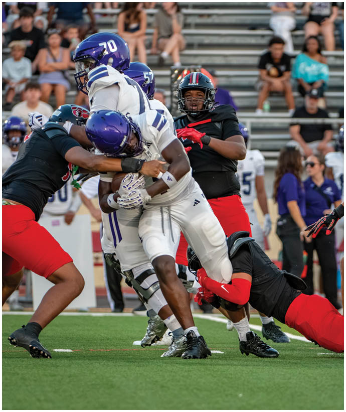 Two football players in black and red uniforms are tackling a player in a white uniform on a green field with spectators in the background. Description generated by AI