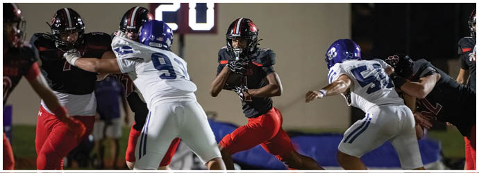 Adults wearing football uniforms are engaged in a game on a field with one player in black and red running while others in white and blue attempt to block or tackle. Description generated by AI