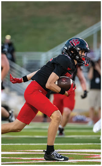 A person wearing a black football jersey and red pants is holding a football while running on a field. Description generated by AI