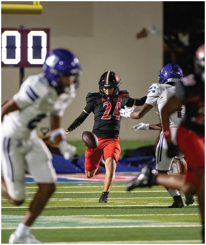 A person wearing a black and red football uniform is kicking a football while two other people in white and purple uniforms are nearby on a football field. Description generated by AI