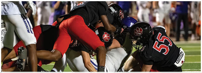 Several football players in black and red uniforms are tackling a player in a white uniform on a green field during a game. Description generated by AI