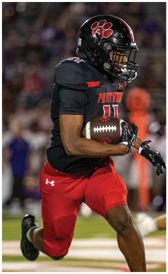 A person wearing a black football jersey with red lettering and red pants is holding a football while running. Description generated by AI