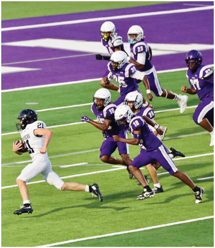 A person in a white sports uniform is running with a football while several people in purple uniforms chase them on a football field. Description generated by AI