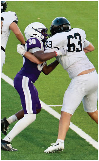 Two people in football uniforms are engaged in a play on a grassy field, with one wearing a white helmet and purple jersey numbered 30 and the other wearing a white jersey numbered 63. Description generated by AI