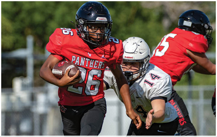 An adult wearing a red Panthers jersey is running with a football while another adult in a white helmet and jersey attempts to tackle them on a football field. Description generated by AI