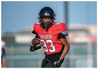 A person wearing a red football jersey with the number 83 and a black helmet is running while holding a football on a field. Description generated by AI
