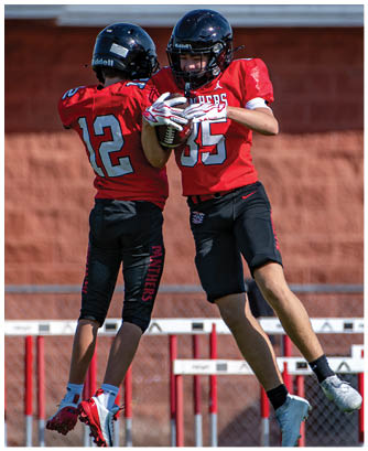 Two people wearing red football jerseys and black helmets are engaged in a play, with one holding a football securely. Description generated by AI