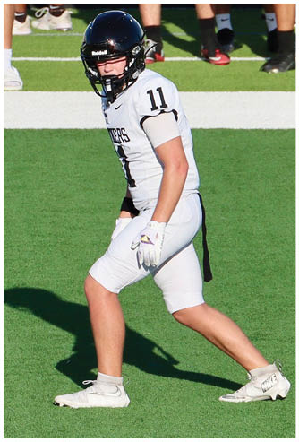 A person wearing a white football uniform with the number 11 and a black helmet is standing on a green field in a ready position. Description generated by AI