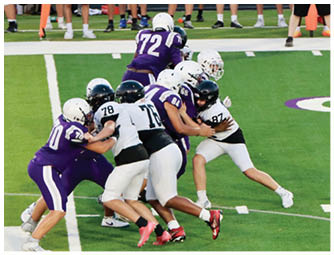 Several people wearing football helmets and uniforms in purple and white are engaged in a play on a green football field. Description generated by AI